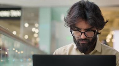 Focused adult ethnic Arabian Indian businessman in glasses typing on computer working indoors. Serious male manager man executive work with laptop analyzing business digital tech data typing online