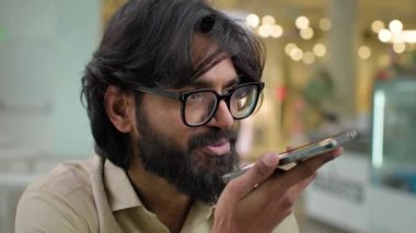 Indian Arabian man in glasses recording voice recognition message on speakerphone. Bearded businessman in eyeglasses guy using virtual assistant app sets reminder on phone digital AI mobile assistance