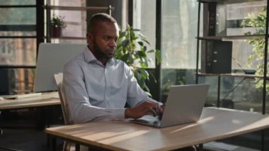 Focused adult senior businessman mature entrepreneur African American ethnic man working in office business firm programming on computer keyboard online project typing email sit in sunbeams background