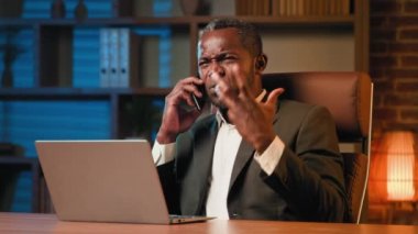 Multitasking african american businessman talking on phone in office near laptop serious upset emotional man speak with employee explain cause problem dissatisfied customer communicate using cellphone