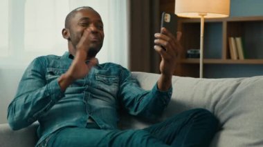 Smiling mature african american man sitting on comfortable sofa in room talk video call using mobile phone waving greet positive male remote communicate with friend by online conference on smartphone