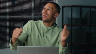 Happy young african american man dancing sitting at workplace joyful businessman celebrating victory rejoicing in success enjoy break at work dance dynamic moving to music celebrate career advancement