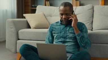 Mature african american businessman freelancer sitting in room floor talking mobile phone working on laptop ethnic man client remote make order in online store using cellphone and computer application