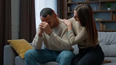 Offended African american man sit on couch upset with bad news health problem Caucasian woman consoling apologize ask forgiveness after quarrel wife touching husband calming support family conflict