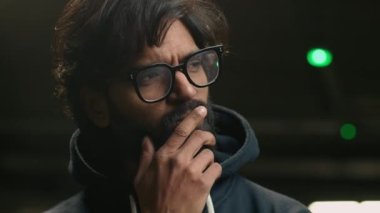 Close up Indian pensive man in eyeglasses thinking looking to side rubbing chin. Thoughtful Arabian male in glasses stand at dark parking lot pondering business idea searching problem solution think