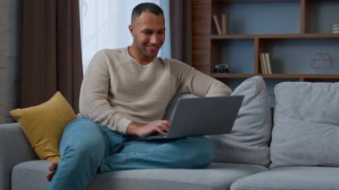 Millennial 30s african american adult man ethnic businessman freelancer sit on couch use pc laptop typing on keyboard distant working chatting online read e-mail e-commerce use home wi-fi networking