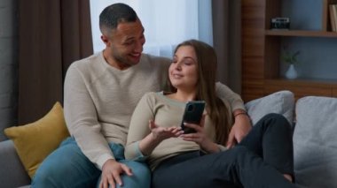 Couple multiracial hispanic woman african american man resting on cozy sofa with smartphone looking at phone screen watch video photo use mobile app talking spend weekend at home with cellphone gadget