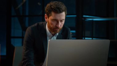 Middle-aged senior businessman Caucasian 40s man adult male employee CEO director manager analyst stand at desk in evening office search business project data online on computer dark night background