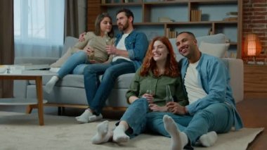Two ethnic couple diverse men women watch television romantic tv film at home family rest on sofa and floor multiracial friends watching comedy movie comfortable leisure in apartment with bottled beer