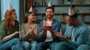 Ethnic diverse women and men in party caps congratulate male caucasian friend presenting surprise cake sing happy birthday song multiracial multiethnic people singing on festive celebration at home