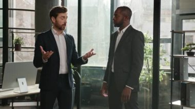 Diverse male coworkers multiracial partners men talking standing in office. Caucasian man manager having discussion with African American employee meeting in company discussing project sharing ideas