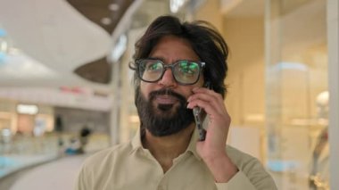 Indian businessman in eyeglasses walk indoors in corporate company office talking phone smile negotiate communication with client with smartphone. Arabian business man speak mobile conversation walk