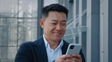 Close up walking in city Asian Korean adult middle-aged 40s businessman smiling happy looking in smartphone chatting browsing in mobile phone boss leader man smile walk work with web online in device