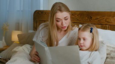 Family learning concept. Caucasian mother help little small cute daughter preschool child girl baby reading book in bed at home bedroom mom with kid read literature fairy tale elementary education