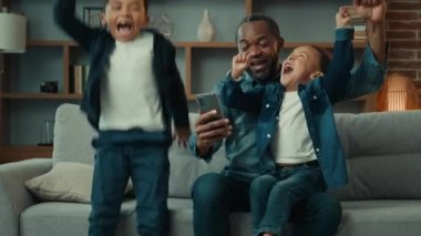 African American man parent father with two boys children little kids siblings play mobile game online win at home couch. Happy dad with sons winning with phone achieve with smartphone scream shout