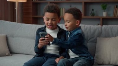 İki küçük Afrikalı Amerikalı kardeş çocuklar cep telefonlu küçük kardeşler internet üzerinden oyun oynuyorlar sohbet ediyorlar evde kanepede oturup cep telefonu uygulaması ile web sayfasını karıştırıyorlar.