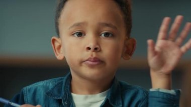 Close up sad serious ethnic school boy child with pen looking at camera waving hand hello hi. Portrait at home calm little kid son pupil schoolboy greeting with arm writing homework lesson education