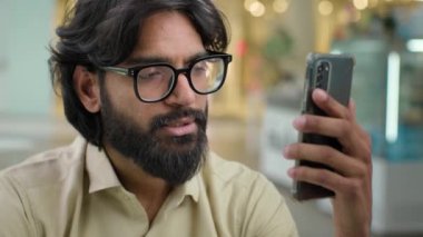 Indian business man Arabian businessman in glasses CEO worker talk mobile phone video call. Serious bearded executive entrepreneur with smartphone app talking distant negotiate virtual communication