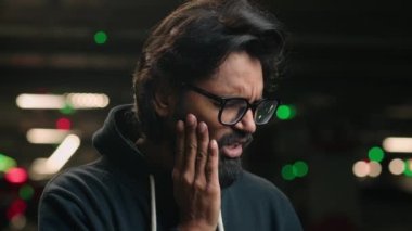 Close up sad ill sick Arabian man in glasses bearded Indian guy with severe toothache. Male at parking lot suffer from strong pain ache painful dental illness teeth problem toothy inflammation of gums