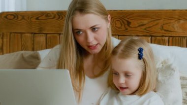 Caucasian mother and cute kid daughter talking using website learning with laptop app at home, mom teach little child girl online shopping make webcam video call looking at computer screen sit in bed