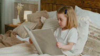 Cute little 5s Caucasian alone child girl daughter looking in book kid preschool baby sitting in bed watching pictures in copybook studying learning read try reading elementary education in bedroom