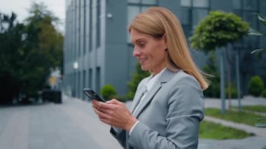 Caucasian businesswoman chatting on internet on mobile phone standing outdoors happy smiling attractive young successful woman using cellphone typing email message communicate by sms side profile view