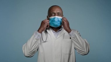 Olgun Afro-Amerikan erkek doktor mavi arka plan stüdyosunda koruyucu maskeyle duruyor. Solunum cihazını çıkarıyor. Derin nefes alıp veriyor.