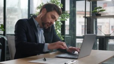 Busy successful caucasian adult businessman in office typing on laptop talking to client on mobile phone writing notes explain condition multitasking man manager communicate on cellphone in workplace