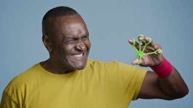 Close-up mature african american athletic man sportsman standing in blue background studio with tense expression doing exercise using sports equipment expander hand trainer for fingers sport training