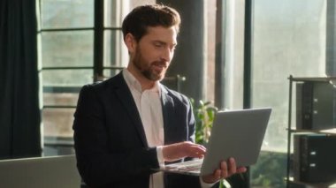 Successful caucasian businessman typing on laptop working on Internet in office in morning sunshine adult happy man leader boss professional manager model smiling looking camera posing at workplace