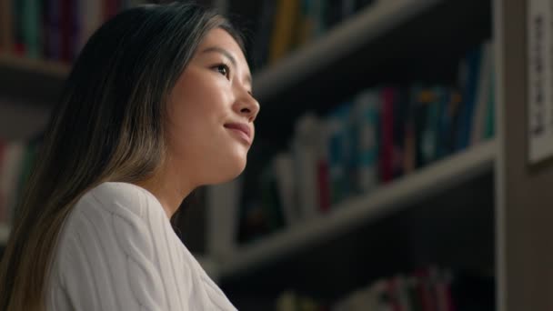 大学図書館の書棚で熟考している若い韓国人女性学生検索で右の教科書に集中した女の子書店で読書する文学を選択する本棚から本を選択 — ストック動画