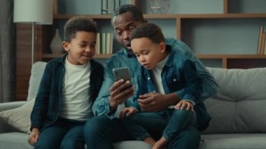 African American family adult father with two school kids children boys kids sons on sofa use funny smartphone apps amazed having fun with technology together look at phone screen video call at home