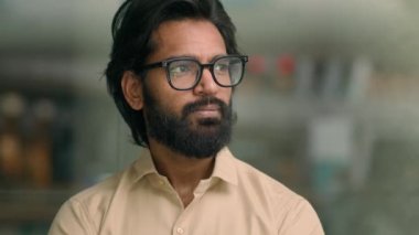 Medium portrait handsome bearded Indian entrepreneur in glasses standing in creative office look to side turning looking at camera. Thoughtful Arabian man businessman contemplating e-business ideas