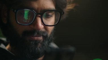 Close up male in eyeglasses Indian bearded businessman Arabian guy texting with smartphone man looking at phone screen green light reflection in glasses using app standing in dark parking lot at night