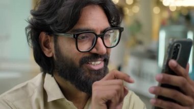 Smiling millennial Indian bearded guy businessman sit in cafe office worker in eyeglasses looking at smartphone screen using mobile phone app scrolling browsing in internet. Arabian man buying online