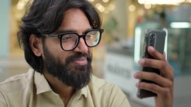 Indian bearded businessman in glasses talk video call on smartphone Arabian guy man executive entrepreneur business specialist talking with mobile phone conference virtual chat indoor greeting hand