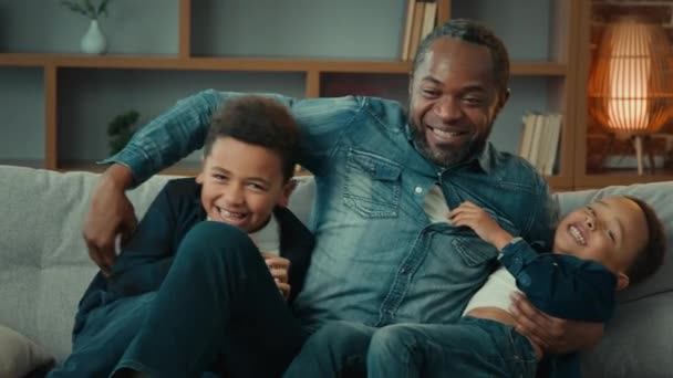 愛情アフリカ系アメリカ人家族お父さんTickling二つの小さなかわいい子供兄弟子供息子抱擁抱っこをします楽しい笑いを持っていますソファで一緒に小さな男の子と一緒に家で遊ぶ — ストック動画