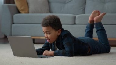 Afrikalı Amerikalı çocuk etnik okul öğrencisi evde internetten çalışıyor ve e-öğrenme adı verilen internet dersi videosunu izliyor. Dizüstü bilgisayarı olan küçük çocuk oturma odasında yerde oyun oynuyor.