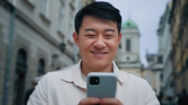 Kapalı çekimde gülümseyen Asyalı turist cep telefonuyla şehirde duruyor. Tatil tatili için cep telefonlu seyahat biletlerini elektronik rehberle kullanıyor.