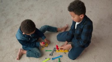 Top view two little siblings boys African American friends kids schoolboys children play game sit on carpet in living room at home playing with plastic toys repair tools hummer screwdriver spanner