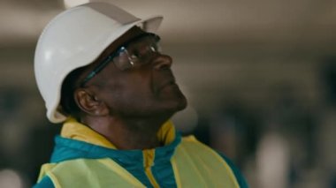 Close-up mature experienced man builder engineer developer inspector in uniform protective helmet goggles inspecting construction african american male architect think about project examines building