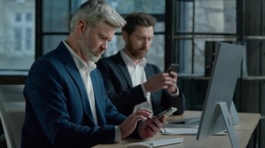 Two caucasian men businessmen business colleagues chat on Internet use mobile phones during break at office check social network on cellphone mature male leader entrepreneur browsing email on phone