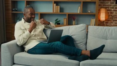 Excited happy mature african american man looking laptop celebrating internet victory lying on couch at home ethnic male winner win online lottery enjoy success read good news in email feel motivated