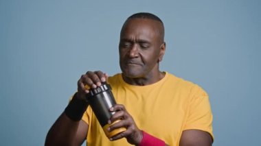 Olgun Afro-Amerikan erkek sporcu mavi arka plandaki stüdyoda sallanıyor. Spor sallama şişesini sallıyor. Enerji kokteyli erkek sporcusu protein beslenme içeceği içiyor.