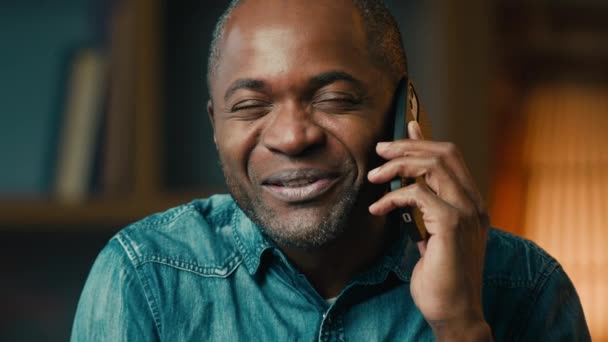 携帯電話で話す幸せな成熟したアフリカ系アメリカ人男性屋内で笑顔の民族男性リモート通信友人とスマートフォンの応答を使用してモバイルコール笑いは電話で話す良いニュースを議論 — ストック動画