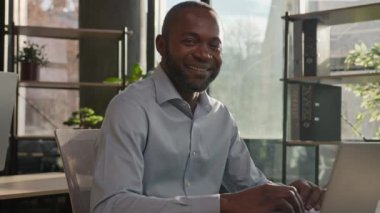 İş portresi Afro-Amerikalı orta yaşlı olgun erkek işadamı güneşli bir ofiste poz veriyor dizüstü bilgisayarla oturuyor ve üst düzey yönetici menajeri kameraya bakıyor.