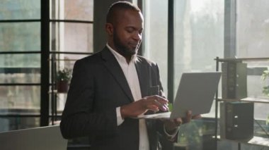 Senior mature businessman African American ethnic man middle-aged male worker develop business project online on computer typing on keyboard stand indoors office with laptop looking at camera smiling