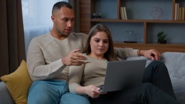 Afrikalı Latin çift dizüstü bilgisayarlı kadın ve erkeğe bakıyor kanepe üzerinde dinleniyor film izleyen farklı aile bireyleri bilgisayar aygıtı kullanıyor e-servis alışverişi yapıyor internette haber alışverişi yapıyor.