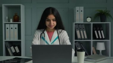 Happy positive woman doctor arabian indian ethnic therapist physician working with laptop looking at camera smiling show okay hand gesture recommend good result approve health care medical insurance