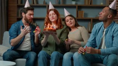 Friends sing happy birthday song to woman congratulate girl at home. Diverse ethnic teammates women and men in party caps presenting festive cake with candles celebrating birth event blowing whistles